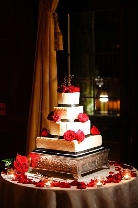 a white wedding cake decorated with black ribbons and red roses on top