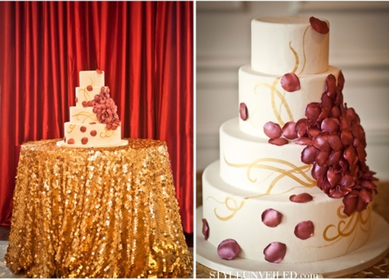 A white wedding cake decorated with burgundy petals, gold patterns served on a dessert table covered with a gold sequin tablecloth