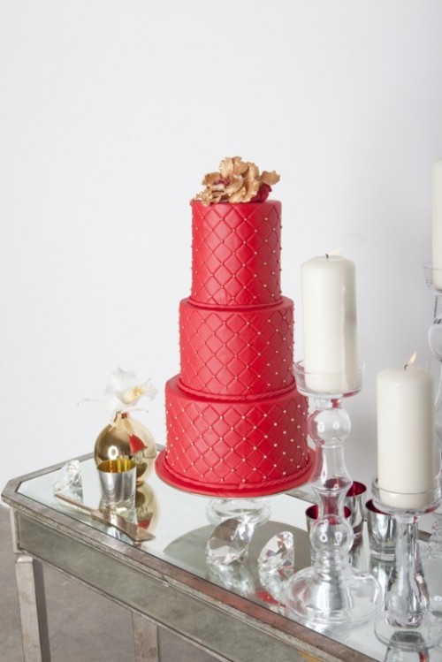 a red patterned wedding cake with gold beads and a large gold sugar flower on top