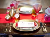 a red table runner, red glasses, gold vases with pink blooms, gold cutlery and a pink rose in a bottle