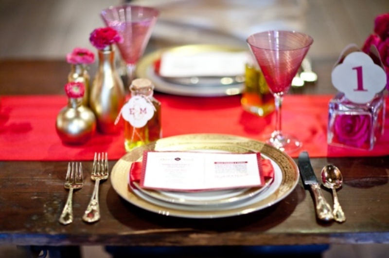 A red table runner, red glasses, gold vases with pink blooms, gold cutlery and a pink rose in a bottle