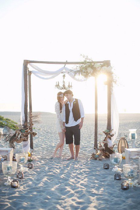 a boho beach wedidng ceremony setting with benches, corals, candles, driftwood and an arch decorated with blooms and airy fabric