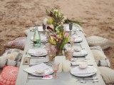 a boho beach wedding reception with a low picnic table, colorful pillows and glasses, bright blooms and greenery