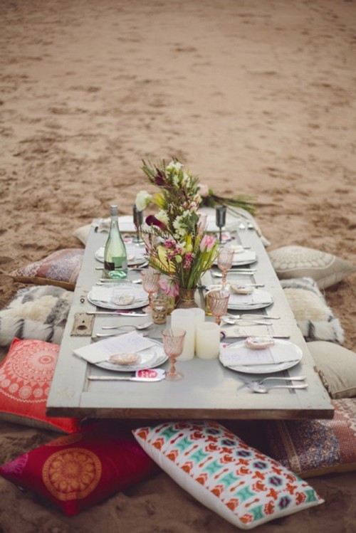 a boho beach wedding reception with a low picnic table, colorful pillows and glasses, bright blooms and greenery