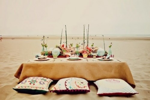 a bright boho beach place setting with an ocher tablecloth, floats, bright blooms and pillows