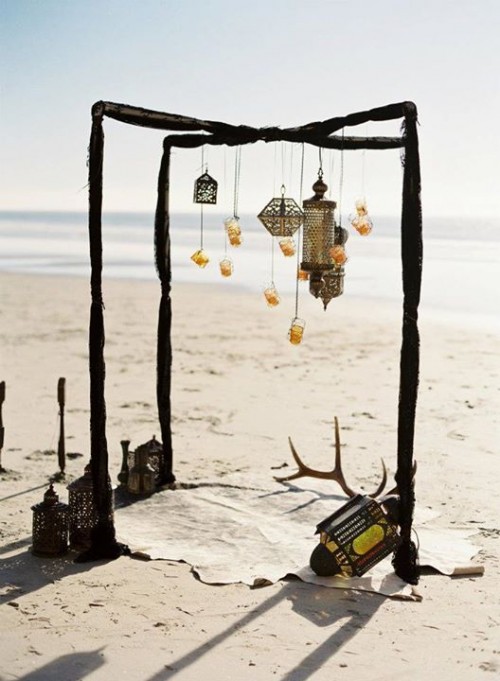 a boho beach wedding ceremony arch done with dark fabric, lanterns and candle lanterns hanging down plus a boho rug