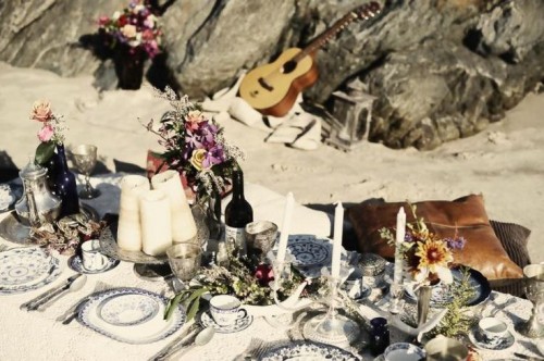 a boho beach table setting with printed plates, candles, bright blooms and various pillows