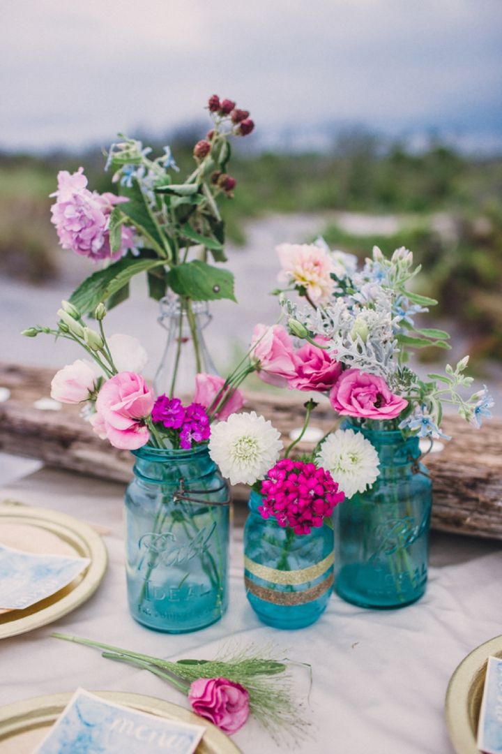 a bright boho beach wedding centerpiece with blue jars and bright blooms and driftwood with candles