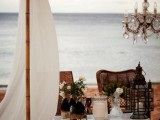 an elegant boho beach wedding picnic setting with a teepee, a crystal chandelier, wicker furniture, a low table and candle lanterns