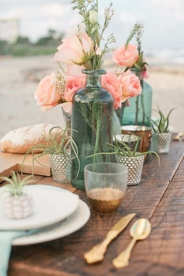 a boho beach wedding centerpiece with candle lanterns, air plants in holders, coral pink blooms and greenery in turquoise bottles