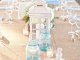 a neutral beach tablescape with a neutral tablecloth, a white candle lantern, blue candle holders and a white hydrangea centerpiece