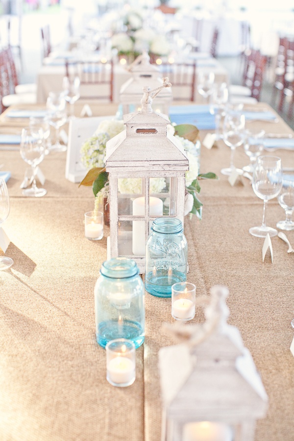 A neutral beach tablescape with a neutral tablecloth, a white candle lantern, blue candle holders and a white hydrangea centerpiece