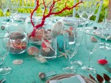 a colorful beach wedding table with a turquoise tablecloth, red corals and seashells, sand and bright turquoise menus