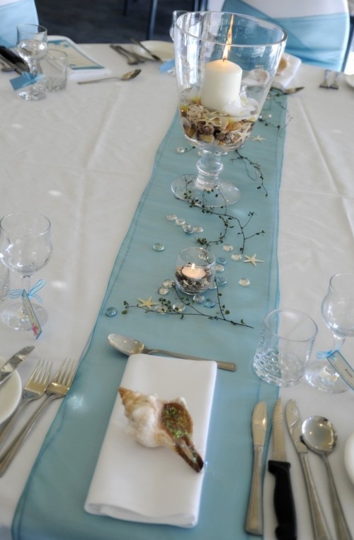 a beach tablescape with a blue runner, twigs, starfish, a glass with seashells and candles plus neutral napkins