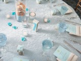 a light blue wedding tablescape with a tablecloth with a sparkle, forsted glass plates and blue menus plus a centerpiece of a tall glass with sand, seashells and a starfish