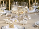a neutral wedding tablescape with a tablecloth, seashells, white blooms, neutral napkins and mini sand castles as favors