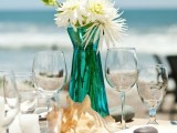 a beach wedding table with an oversized shell, a turquoise vase with neutral blooms, white candles and pebbles on the plates