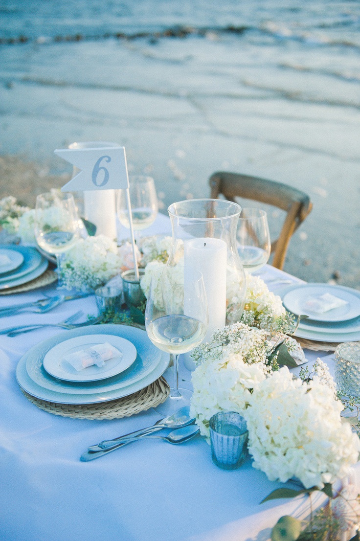 A beautiful light blue and neutral beach wedding tablescape with neutral blooms, blue plates and glasses and woven placemats