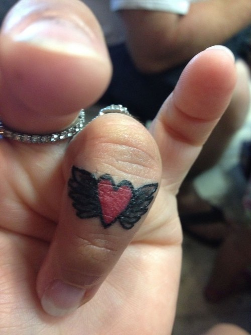 a red heart and wing tattoo on the top part of the ring finger is a bold and lovely idea to rock