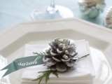 a white and silver place setting with a white napkin, a snowy pinecone, beads and ribbons plus evergreens