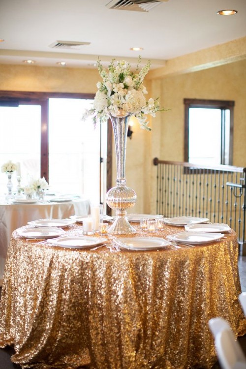 a shiny and sparkling wedding reception table with a gold sequin tablecloth, a tall white floral centerpiece and white porcelain