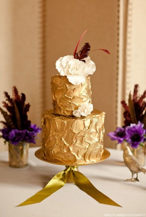 a textural gold wedding cake with white blooms and burgundy feathers on top is a whimsical and cool idea for a refined wedding