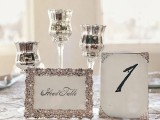 a silver wedding centerpiece of silver candleholders and an embellished card with calligraphy and a table number