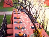 a stylish rustic Halloween wedding tablescape with a red tablecloth, black napkins, pumpkins, a planter with branches and bright blooms and leaves