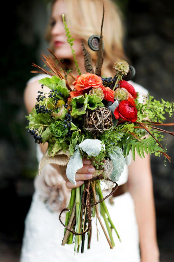A bright woodland boho wedding bouquet of fern, greenery, berries, bright blooms, feathers, and lotus pieces