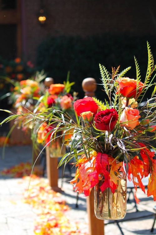bright floral arrangement of red, orange roses, greenery and bold fall leaves is a cool idea to style a fall wedding aisle