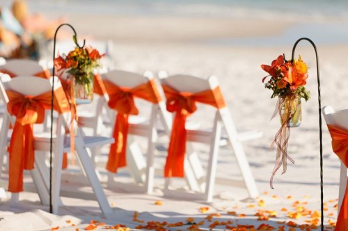 a fall beach wedding aisle with bright orange and yellow blooms and greenery, with long ribbons and orange petals right on the ground