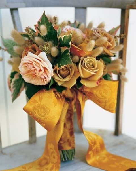 a bright warm-colored wedding bouquet of yellow and blush blooms, large leaves and bunny tails plus a gold yellow printed ribbon bow is cool for a pretty and bold rustic wedding