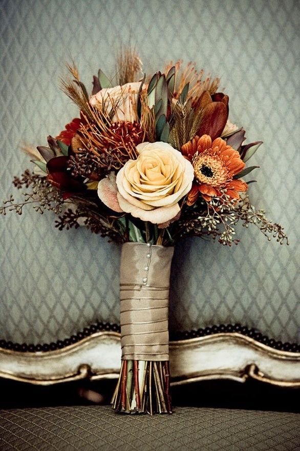 A bold fall boho wedding bouquet of rust, burgundy and neutral blooms, feathers and dark foliage, gilded greenery plus a tan wrap