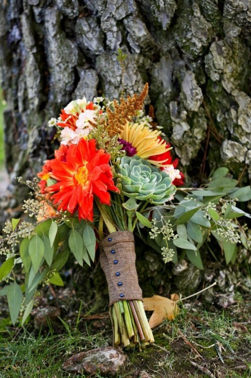 a bold fall wedding bouquet of orange, red, yellow and white blooms, greenery, grasses and a succulent plus a burlap wrap on buttons is a gorgeous idea for a colorful fall rustic wedding