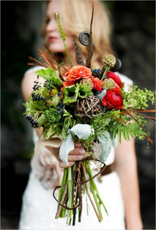 a colorful and textural fall wedding bouquet of greenery, orange and depe red blooms, a vine ball, privet berries, feathers and lotus is a catchy idea for a fall woodland bride