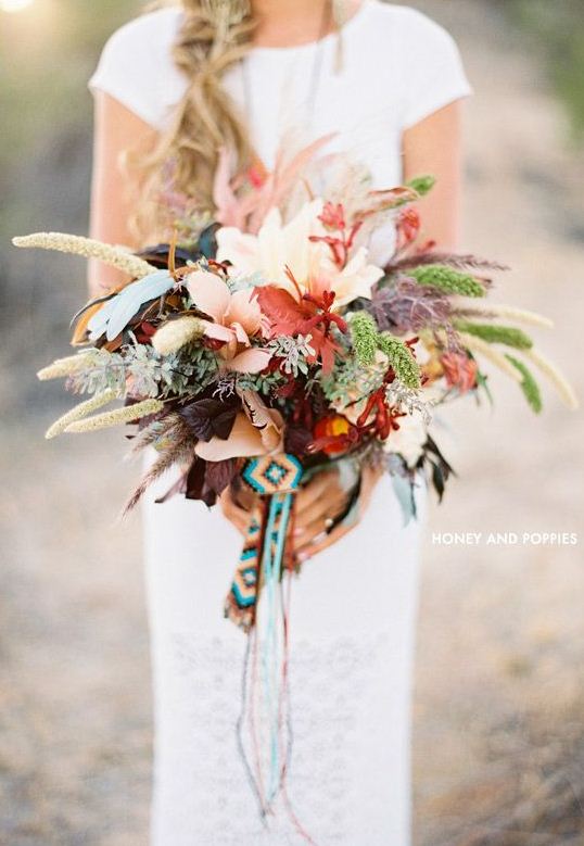 A lush dimensional fall wedding bouquet with greenery, feathers, foliage and bright blooms plus a colorful boho wrap and ribbons is a cool idea for a boho fall bride