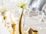a chic and shiny minimalist wedding tablescape with a gold runner, some gold candle holders and a vase with neutral blooms