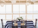 a chic nautical table setting with a striped navy and white tablecloth, dark chairs, a white floral centerpiece and white paper lamps over it