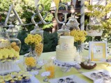 a yellow and grey dessert table with a banner, artworks and yellow flowers, a yellow cake and silver sweets