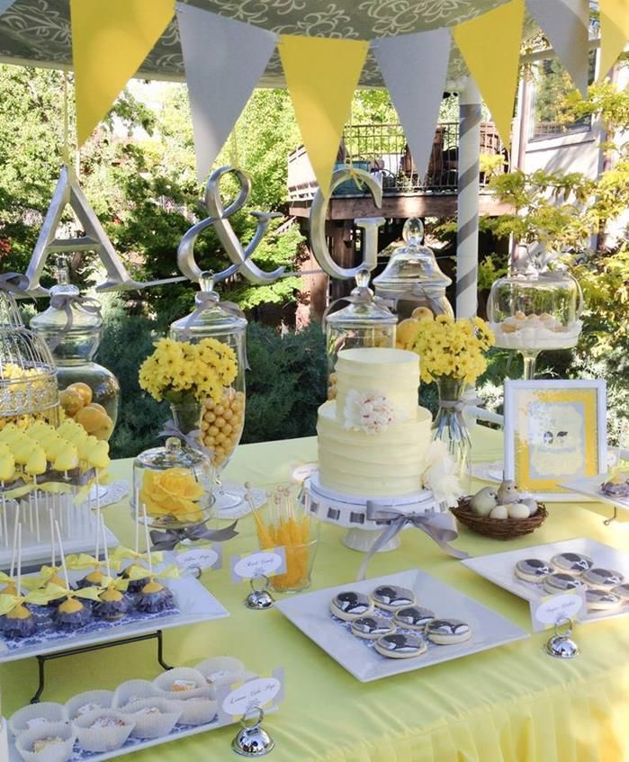 A yellow and grey dessert table with a banner, artworks and yellow flowers, a yellow cake and silver sweets