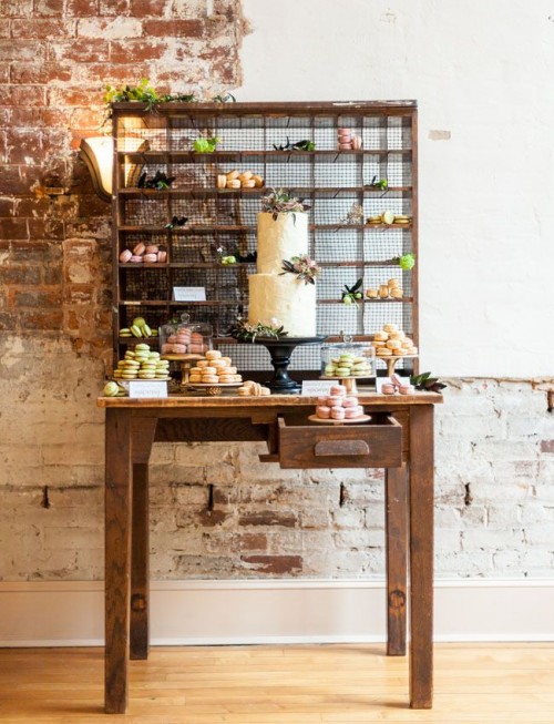 a rustic dessert table with a stand with bright macarons, blooms and greenery and a cake on a chic black stand