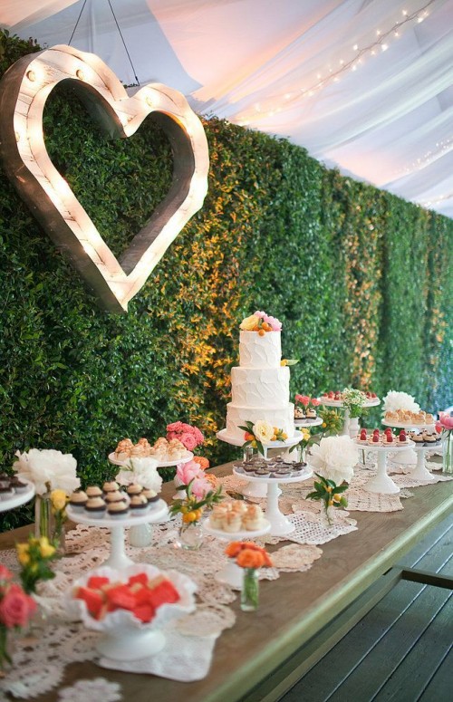 a rustic table with a doliy runner, a green living wall as a backdrop and a marquee heart on the wall