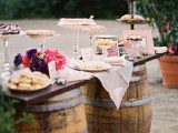 a vineyard wedding dessert table with barrels and bright flowers plus silver stands and glass ones