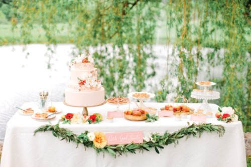 a summer dessert table with greenery and bright flowers and bright desserts and cakes topped with flowers