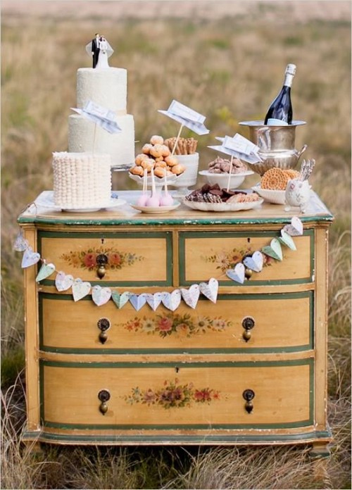 a rustic wedding dessert table with a paper banner, sweets with toppers and some champagne in a bucket
