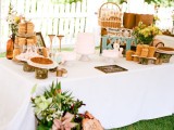 a white dessert table covered with a tablecloth, with pink blooms and greenery, wicker lamps and baskets for holding food