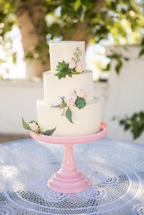 a neutral buttercream wedding cake with blush blooms, succulents and greenery, on a pink stand, is a lovely solution for a modern wedding in spring or summer