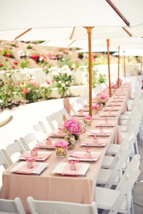 a pastel pink summer wedding tablescape with pink linens, bright blooms, favor boxes and umbrellas over all the tables to prevent excessive sunshine