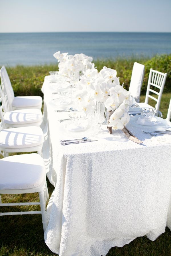 a chic white summer wedding tablescape with a textural tablecloth, lush white orchids and simple linens and cutlery with a sea view