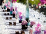 a super bold summer wedding tablescape with purple blooms in blue vases, candles in dark candleholders and all white everything to create a contrast